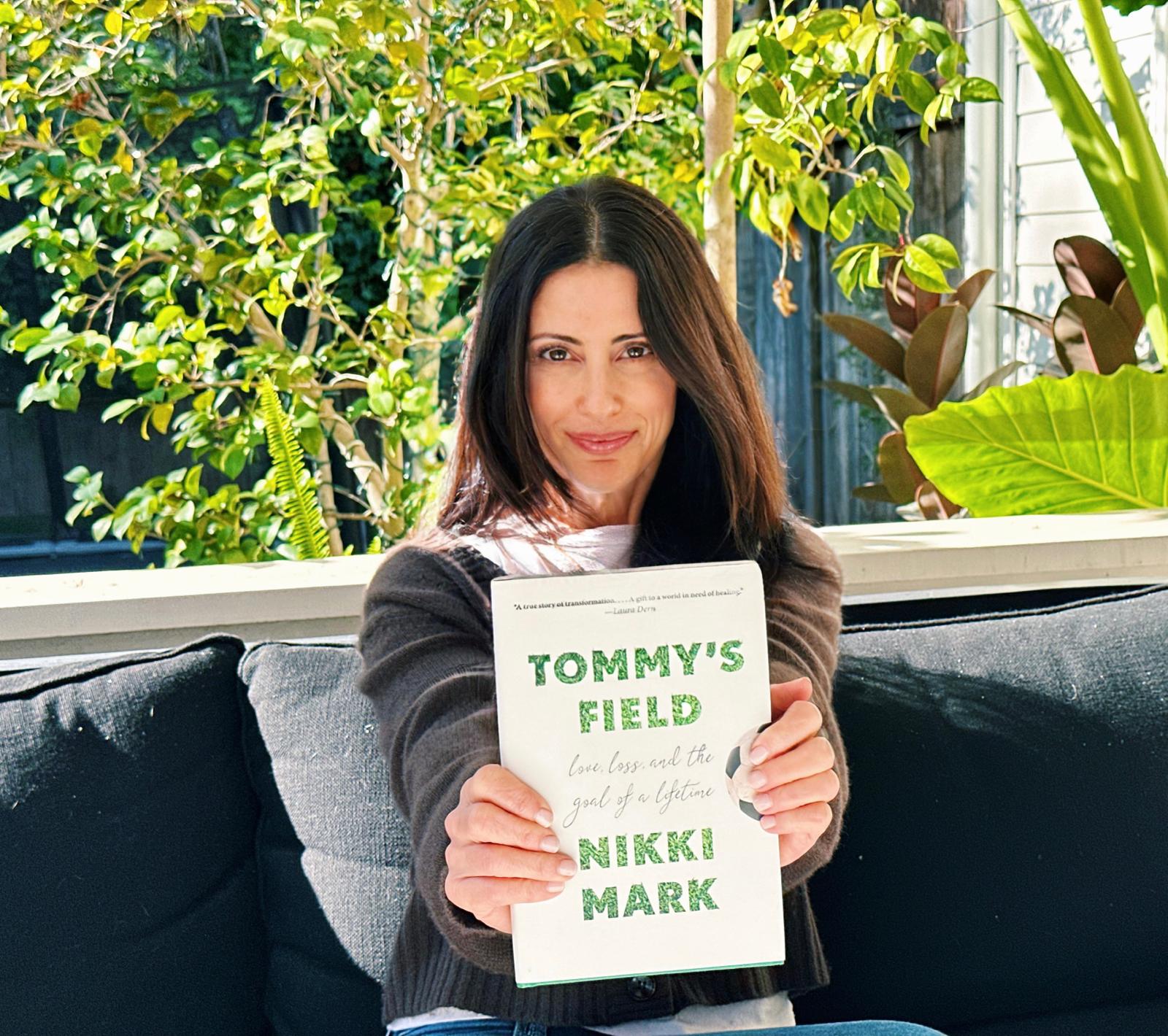 Woman holding a book titled "Tommy's Field" outdoors.