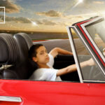 Child driving red convertible on racetrack at sunset.